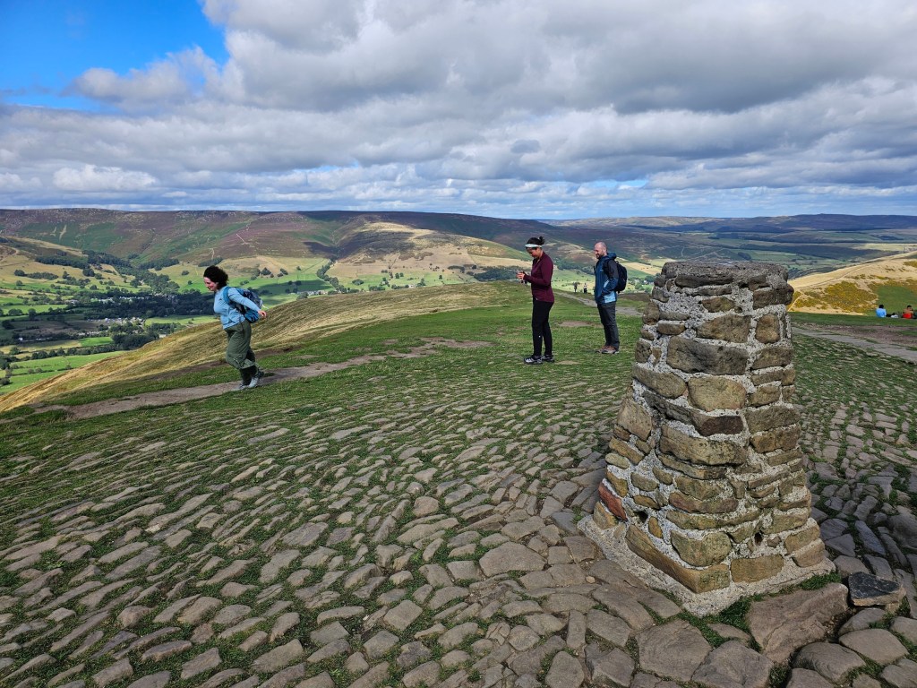 Trig Point on top of Mam Tor. Other hills can be seen in the background. There are a few people seen taking photos.